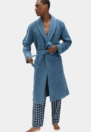Turkusowy szlafrok z miękkiego, teksturowanego materiału z kołnierzem typu szalowego i wiązanym w pasie. Noszony na granatowo-białych, kratkowanych spodniach do piżamy.