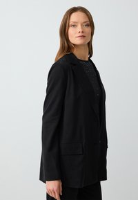 Blazer nero con una vestibilità su misura, realizzato in tessuto morbido. Presenta un colletto a punta, chiusura con un solo bottone e tasche frontali.