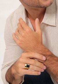 Anello in metallo color oro con un accento rettangolare nero. Presenta un design a strati. Mani esposte su uno sfondo neutro.