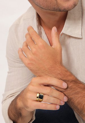 Gold-tone metal ring with a rectangular black accent. Features a layered design. Hands displayed against a neutral background.