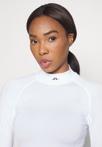 Long-sleeve, high-neck top in white stretch fabric with a black logo on the collar, featuring seamless design and smooth texture.