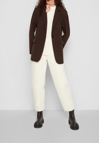 Blazer marrone con rever e tasche, abbinato a un top crema e pantaloni bianchi a 3/4. Stivali con suola spessa neri completano l'outfit.