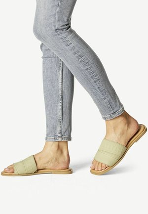 Sandales à glissière vert clair avec deux larges brides en tissu, semelle plate et orteils ouverts, portées avec un jean skinny gris. Présentent un design texturé.