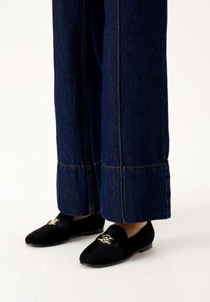 Mocassini in suede neri con un'emblema decorativo dorato sulla parte anteriore, abbinati a pantaloni in denim scuri a gamba larga con cuciture a contrasto.