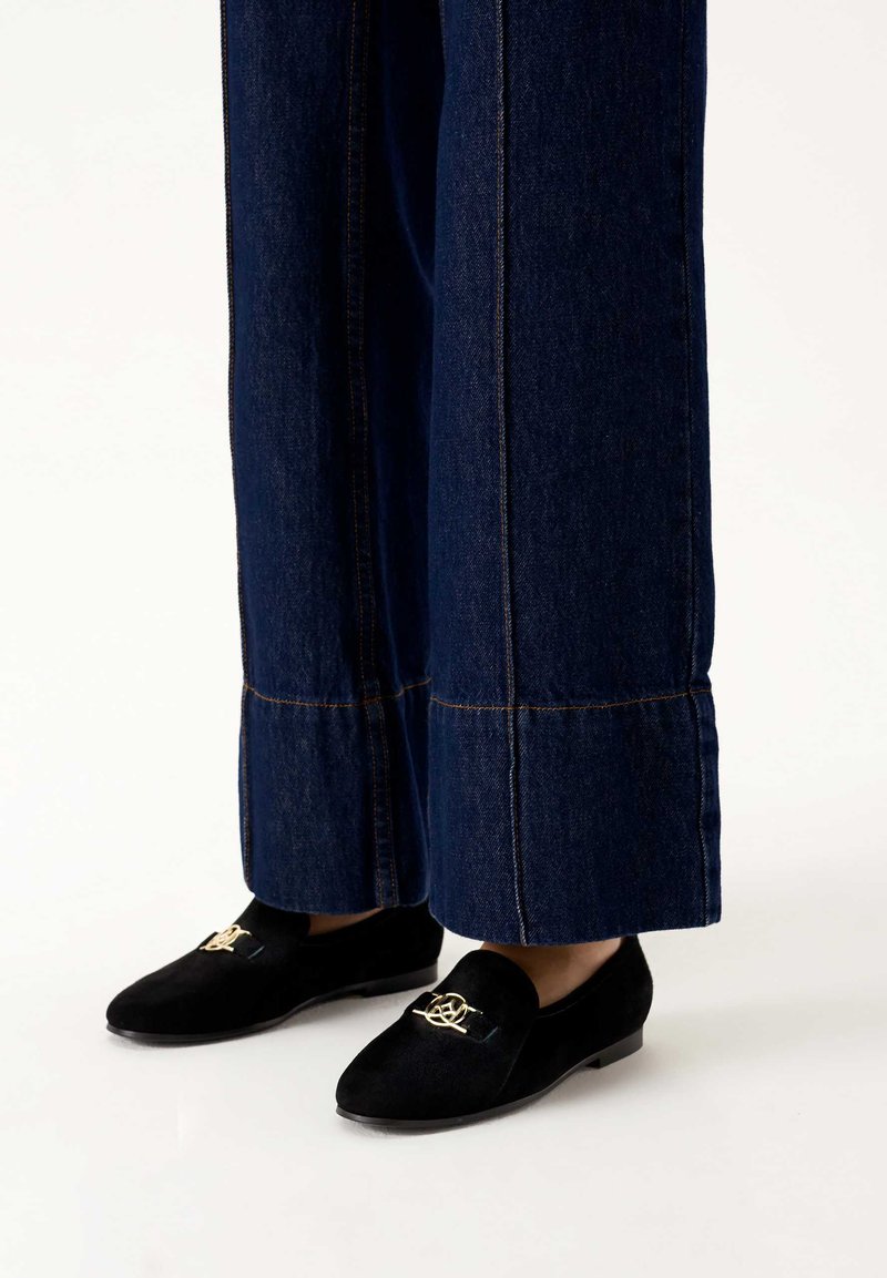 Mocassini in suede neri con un'emblema decorativo dorato sulla parte anteriore, abbinati a pantaloni in denim scuri a gamba larga con cuciture a contrasto.