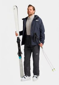 Skiausrüstung umfasst weiße Skier mit schwarzen Bindungen, schwarze Skihosen, eine marineblaue Jacke und weiße Skischuhe. Die Person hält Skistöcke in der Hand.