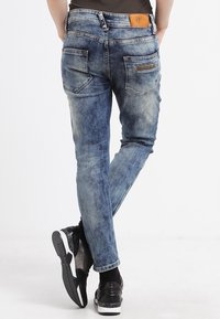 Jeans in denim blu con una finitura sbiadita, cinque tasche e un'etichetta in pelle marrone sul retro. Design slim-fit e texture leggermente usurata.