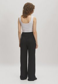 Schwarze, weit geschnittene Hose mit texturierter Oberfläche und elastischem Bund. Trägt ein enges weißes Tanktop, von hinten gesehen.