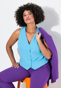 Gilet senza maniche azzurro chiaro con cinque bottoni, abbinato a pantaloni viola. La modella tiene una giacca viola abbinata sopra la spalla.