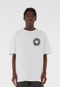 Biela voľná tričko s krátkymi rukávmi, na hrudi s kruhovou čiernou grafikou zobrazujúcou zložitý dizajn a symboly.