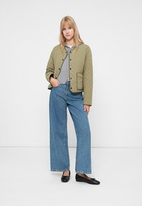 Blondynka w oliwkowej pikowanej kurtce, szarej bluzie z kapturem, szerokich niebieskich dżinsach i czarnych baletkach stoi na białym tle.