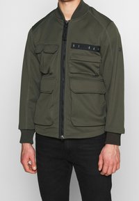 Olivgrön bomberjacka gjord av mjukt, strukturerat tyg som har en framzip, fyra framfickor, ribbstickade ärmslut och dekorativ sömnad.