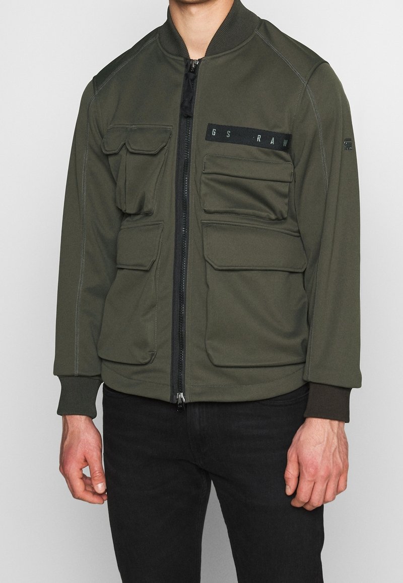 Olivgrön bomberjacka gjord av mjukt, strukturerat tyg som har en framzip, fyra framfickor, ribbstickade ärmslut och dekorativ sömnad.