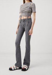 Mujer vistiendo un top corto arrugado gris, jeans de campana grises de cintura alta y sandalias blancas de tiras, de pie contra un fondo liso.