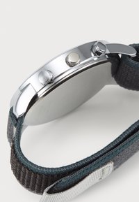 Orologio argento con cassa tondeggiante, cinturino in tessuto grigio, due pulsanti e corona texturizzata; design minimalista e finitura liscia.