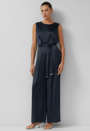 Kobieta w granatowym, satynowym kombinezonie bez rękawów z szerokimi nogawkami i paskiem w talii, w połączeniu ze srebrnymi sandałami na obcasie z paskami.