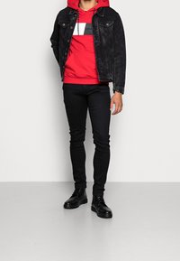 Giacca di jeans nera sopra un hoodie rosso con design grafico, abbinata a jeans neri slim-fit e stivali neri alla caviglia.
