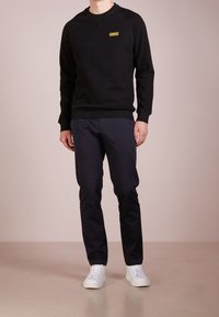 Black sweatshirt with a crew neckline, featuring a yellow logo patch. Paired with navy trousers and white sneakers. Smooth fabric texture.