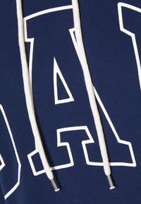 Sudadera azul marino con letras blancas grandes y cordones color crema con terminales metálicos, hecha de una tela suave con un acabado texturizado.
