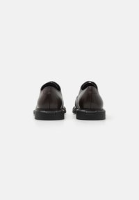 Une paire de chaussures de ville en cuir marron foncé avec des semelles noires, vue de l'arrière sur un fond blanc uni.