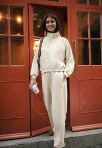 Beez-sweater poolsariga poole tõmbega ja brodüüritud "SELF." logoga, koos sobivate trenni-pükstega; sile tekstuur; seismas punaste uste ees.