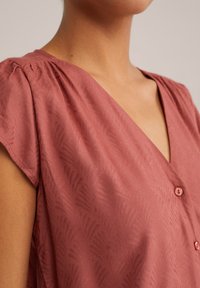 Blusa rosa con scollatura a V, maniche corte a palloncino e una sottile texture a motivo foglia. Include bottoni abbinati lungo la parte anteriore.