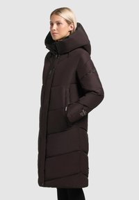 Garš, tumši brūns pūkains mētelis ar lielu kapuci, quilted dizains, ar rāvējslēdzēju kabatām un regulējamām auklām. Gluda tekstūra un eleganta silueta.