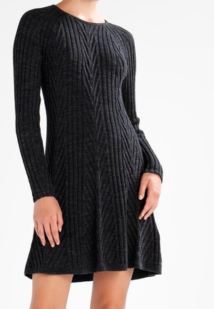 Een vrouw toont een ribbelige, lange mouwen, donkergrijze jurk, waarbij ze de aansluitende bodice en de uitlopende rok laat zien tegen een eenvoudige witte achtergrond.