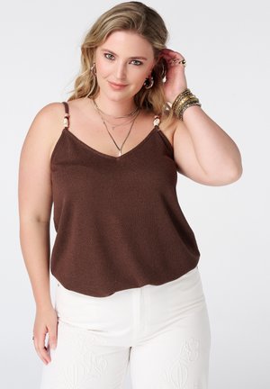 Vrouw met blond haar die een bruine mouwloze top en een witte broek met bloemenborduursel draagt, gecombineerd met armbanden en kettingen.