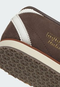 Brun mockasneaker med vita läderdetaljer och zickzack-sömmar. Guldfärgad logga på sidan och en strukturerad gummisula.