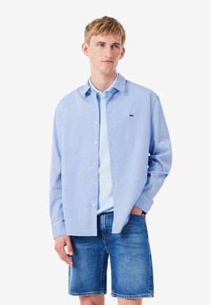 Joven con una camisa de cuadros azul claro abotonada sobre una camiseta polo azul claro y pantalones cortos vaqueros azules, de pie con una mano en el bolsillo.