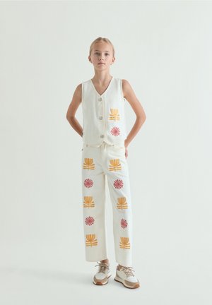 Niña de pie con las manos detrás de la espalda, vistiendo un chaleco blanco y pantalones decorados con estampados de flores naranjas y rosas, y zapatillas deportivas beige.