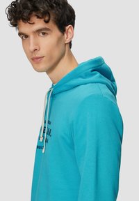 Türkiser Hoodie mit Kordelzug, versehen mit einem gedruckten schwarzen Logo auf der Vorderseite. Der Stoff wirkt weich und hat ein lässiges Design.
