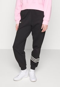 Osoba v černých joggingových kalhotách Adidas s bílými pruhy u nohavic, zkráceném růžovém svetru, bílých ponožkách a bílých teniskách stojí na jednoduché podlaze.
