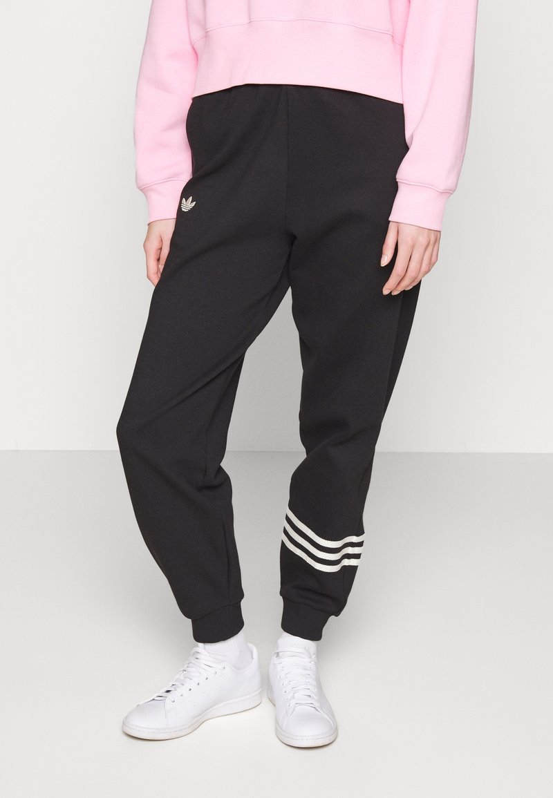 Osoba v černých joggingových kalhotách Adidas s bílými pruhy u nohavic, zkráceném růžovém svetru, bílých ponožkách a bílých teniskách stojí na jednoduché podlaze.