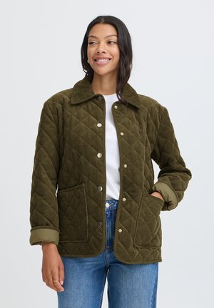 Femme souriante portant une veste matelassée vert olive avec des boutons argentés, une chemise blanche et un jean bleu, debout devant un fond clair uni.