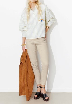 Femme portant une chemise rayée boutonnée et un pantalon beige, tenant une veste en daim marron, debout contre un mur blanc, portant des sandales plateformes noires.