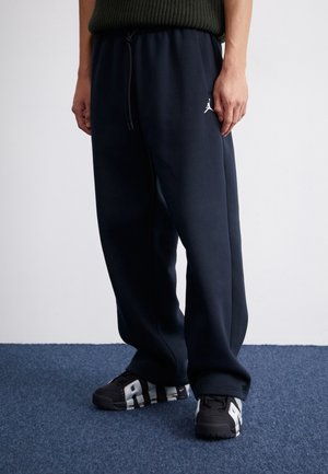 Osoba ubrana w czarne dresy z białym logo Jumpman na udzie oraz czarno-białe buty sportowe Nike stoi na niebieskim dywanie.