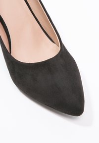 Scarpa da donna in suede nero con punta affusolata e interno in pelle beige, esposta su uno sfondo bianco.