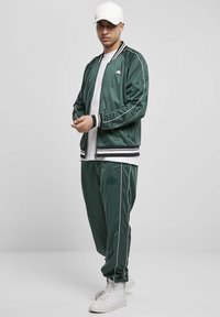 Giacca bomber in satin verde con rifiniture a strisce nere e bianche, abbinata a pantaloni jogger coordinati con un logo. Indossata con sneakers bianche e un cappellino.
