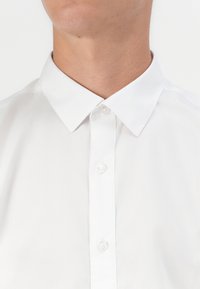 Gros plan d'une personne portant une chemise blanche boutonnée avec un col pointu sur un fond clair uni.