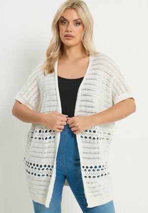 Mujer rubia con un cárdigan blanco de punto abierto sobre una blusa negra y vaqueros azules, mirando al frente con las manos sujetando los bordes del cárdigan.