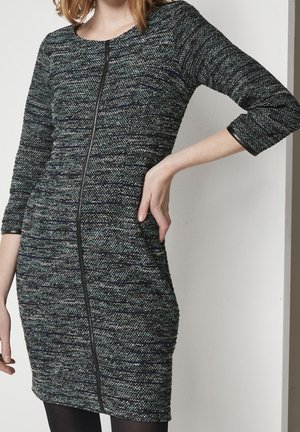 Femme portant une robe ajustée à texture, longueur genou, manches trois-quarts, avec un détail de fermeture éclair noire verticale à l'avant.