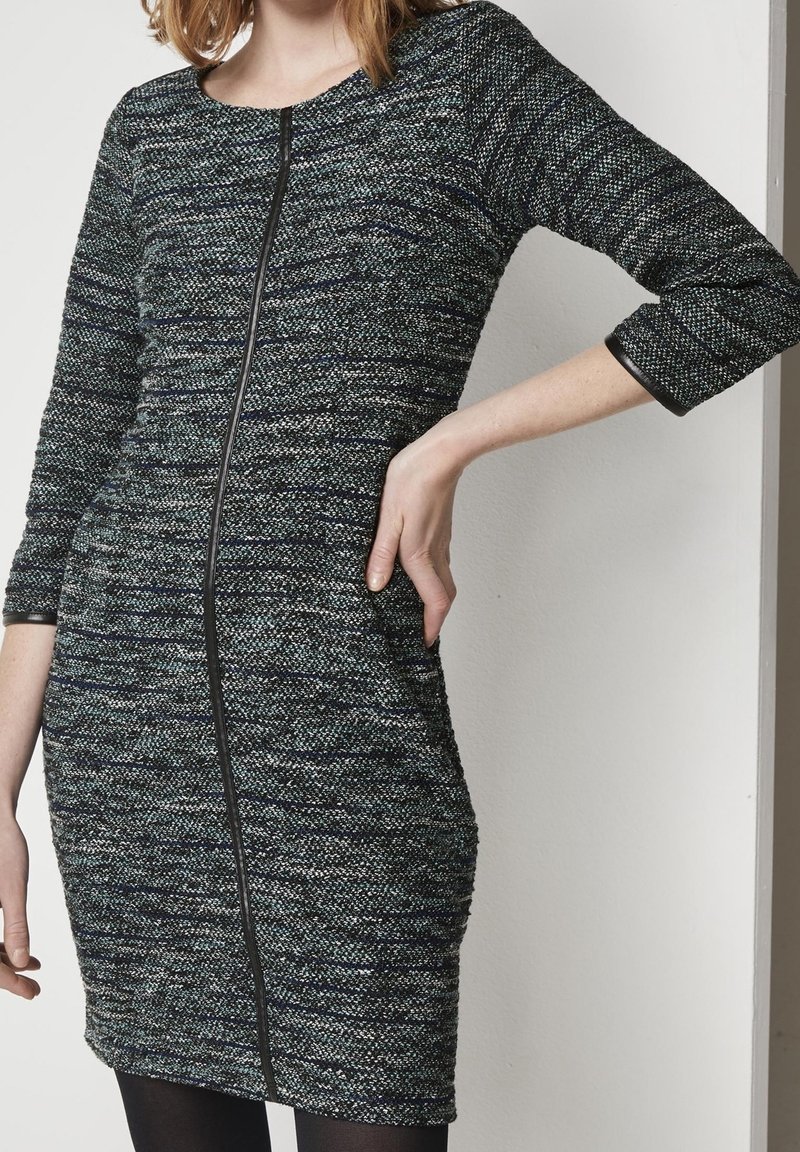 Femme portant une robe ajustée à texture, longueur genou, manches trois-quarts, avec un détail de fermeture éclair noire verticale à l'avant.