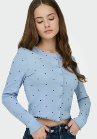 Cardigan bleu clair côtelé avec pois noirs, doté d'un design court, d'un col rond et d'un ourlet à volants. Les boutons avant sont bleus.
