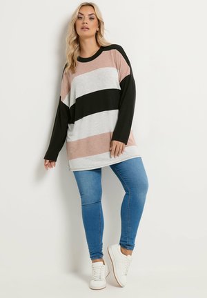 Kvinde iført oversized langærmet sweater med beige, hvide og sorte striber, blå jeans og hvide sneakers, stående mod en hvid væg.