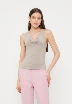 Ung kvinde med langt brunt hår iført guldringeøreringe, ærmeløs beige top og lyserøde bukser mod en hvid baggrund.