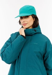 Chaqueta aislante de color azul verdoso con alto cuello, puños interiores negros y bolsillos con cremallera; combinada con una gorra turquesa a juego con visera plana.