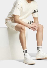 Beige bomullst-shirt och shorts med svarta ränder. Vita sneakers med tre ränder och logotyp, kombinerade med ankelsockor med svarta ränder.
