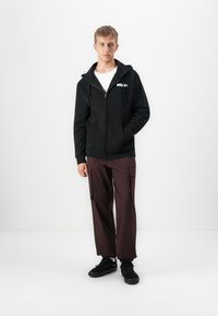 Ung mann iført en svart hettegenser med glidelås, hvit t-skjorte, mørke cargo-bukser og svarte joggesko som står mot en hvit bakgrunn.